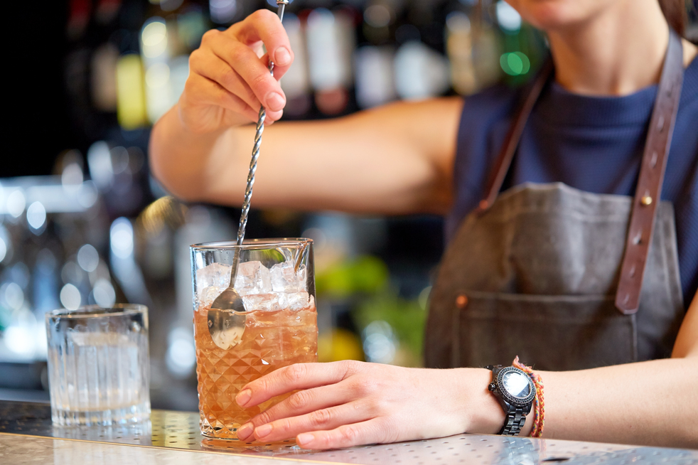 alcohol drinks, people and luxury concept - woman bartender with stirrer and glass preparing cocktail at bar counter