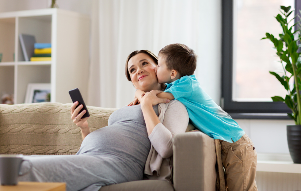 family, emotions and pregnancy concept - little son kissing happy pregnant mother with smartphone at home