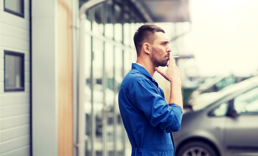 service, repair, maintenance and people concept - auto mechanic smoking cigarette at car shop or garage outdoors